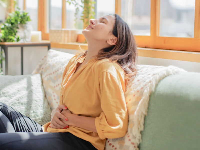 一位身穿黃色上衣的女性，坐在淺綠色的沙發上，閉著眼睛，臉上帶著微笑，雙手輕輕放在腹部。