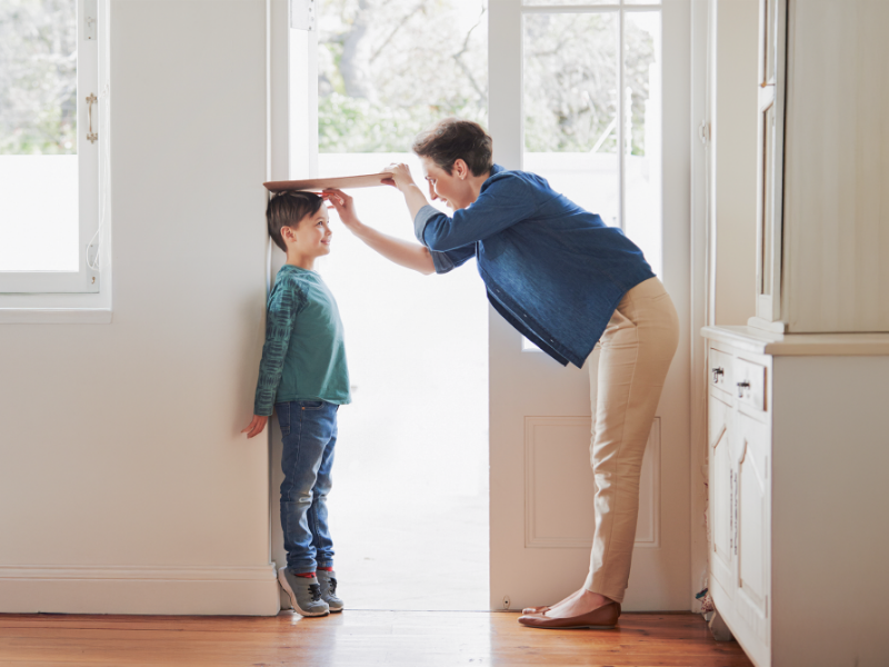 一位年輕的父親正彎下腰，用一塊木板在門框上為身穿綠色上衣的兒子測量身高。兒子面帶微笑，父親也專注於測量。